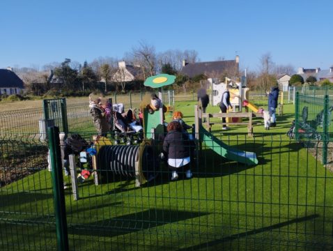 Jeunes enfants jouant dans une aire de jeux sécurisée en plein air pour tout-petits, entourés de leurs accompagnateurs, sur un sol souple en gazon synthétique dans un village résidentiel.