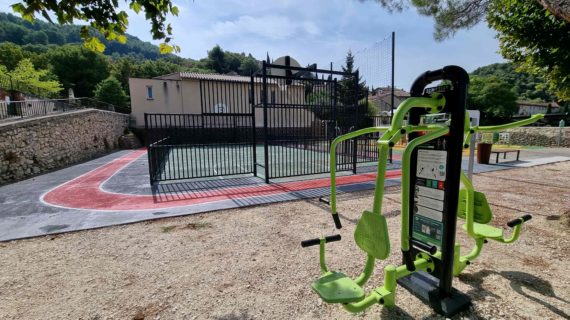 Agrès de musculation extérieur vert face à un city stade clôturé avec piste rouge dans un espace public