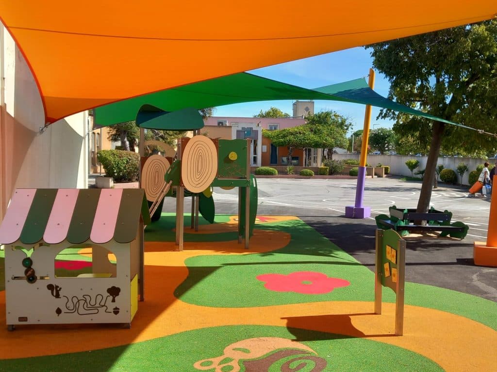 Espace de jeux pour enfants avec cabane, toiles d’ombrage vertes et orange, et sol souple coloré.