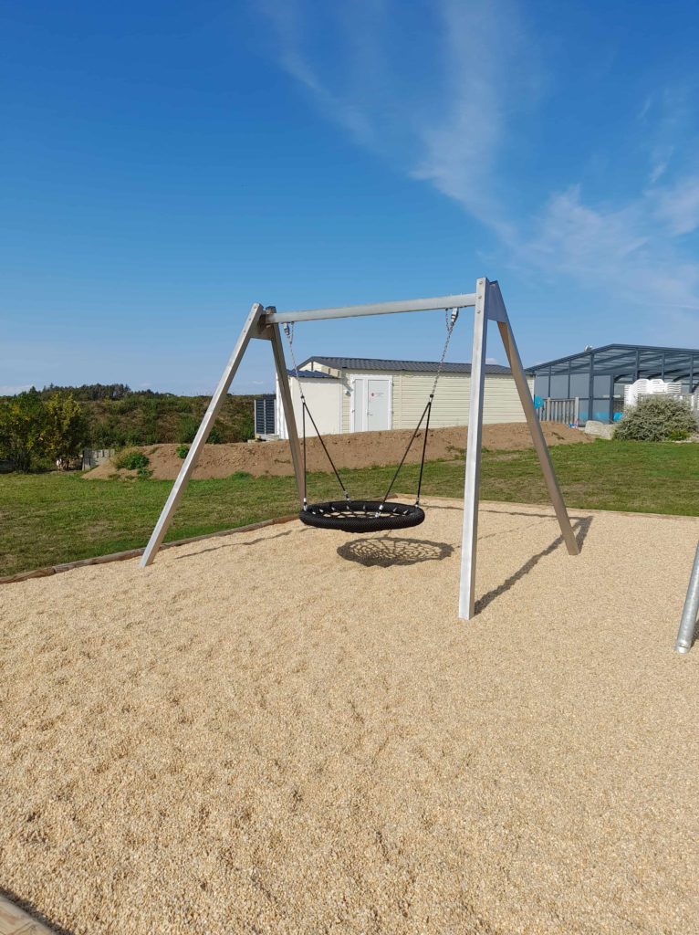 Balançoire nid d'oiseau installée sur une aire de jeux avec sol amortissant en copeaux, dans un espace extérieur naturel.
