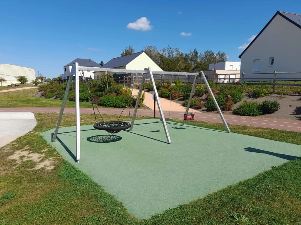 Balançoire nid d'oiseau pour aire de jeux posée sur un sol amortissant en EPDM vert, au cœur d’un espace paysager