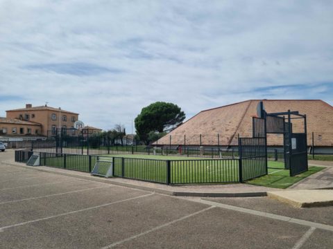 Terrain multisports en extérieur entouré de clôtures noires, avec paniers de basketball et revêtement synthétique, situé près de bâtiments et d’un gymnase.