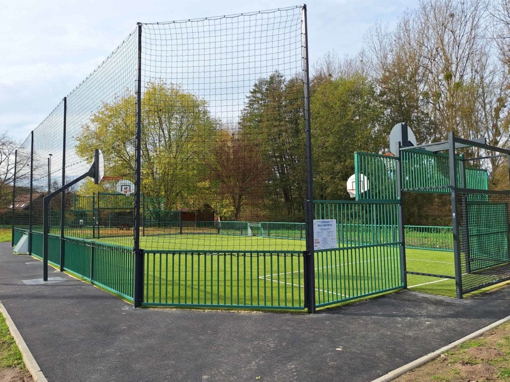 Terrain multisports extérieur avec clôture verte, paniers de basketball et filet pare-ballons, situé dans un espace arboré.