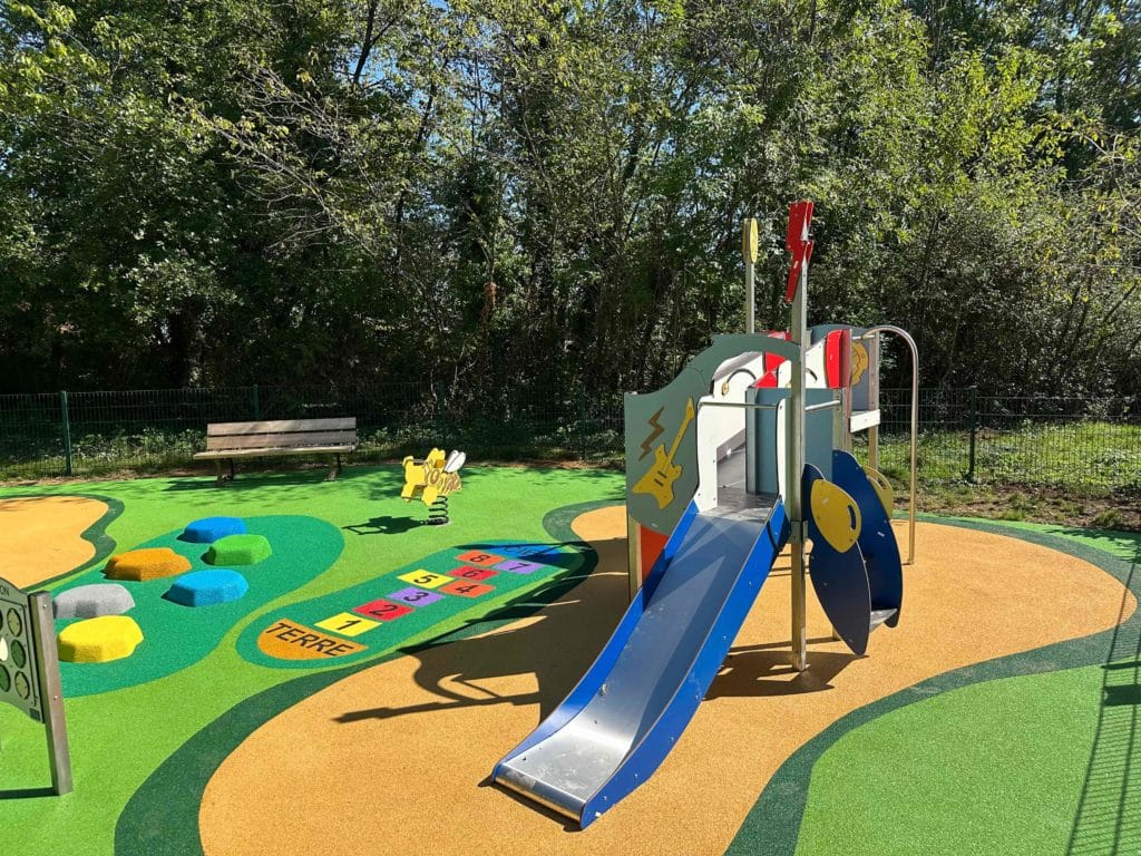 Aire de jeux pour enfants avec un toboggan, un sol souple décoratif aux formes colorées et un parcours ludique comprenant une marelle et des modules en mousse, installée en pleine nature.