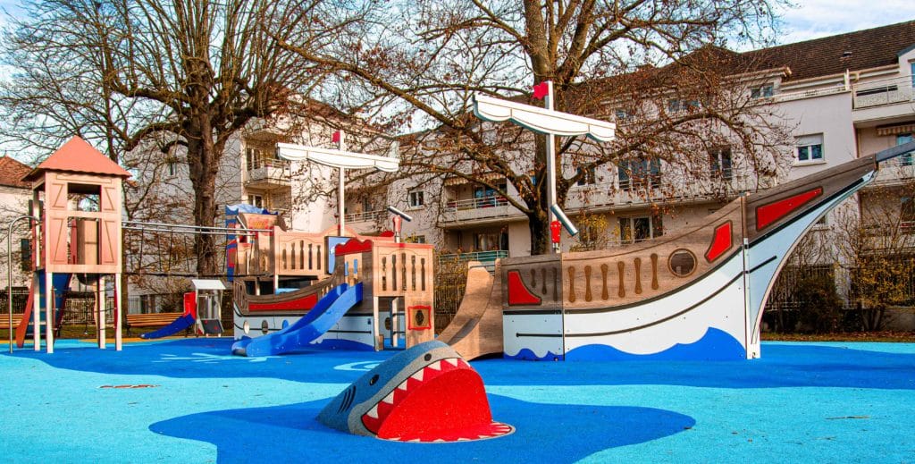 Vue d'ensemble d'une aire de jeux thématique pirate avec bateau, proue de navire, requin en 3D et toboggan sur sol souple amortissant en EPDM