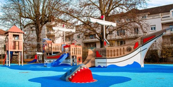 Vue d'ensemble d'une aire de jeux thématique pirate avec bateau, proue de navire, requin en 3D et toboggan sur sol souple amortissant en EPDM