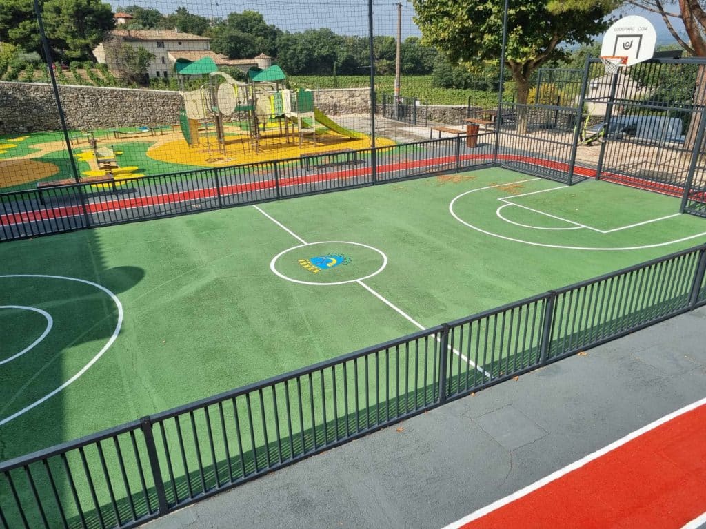 Terrain multisports clôturé avec sol en enrobé, intégrant un marquage sportif complet et le logo de la ville, réalisé à Gigondas, dans le département du Vaucluse.