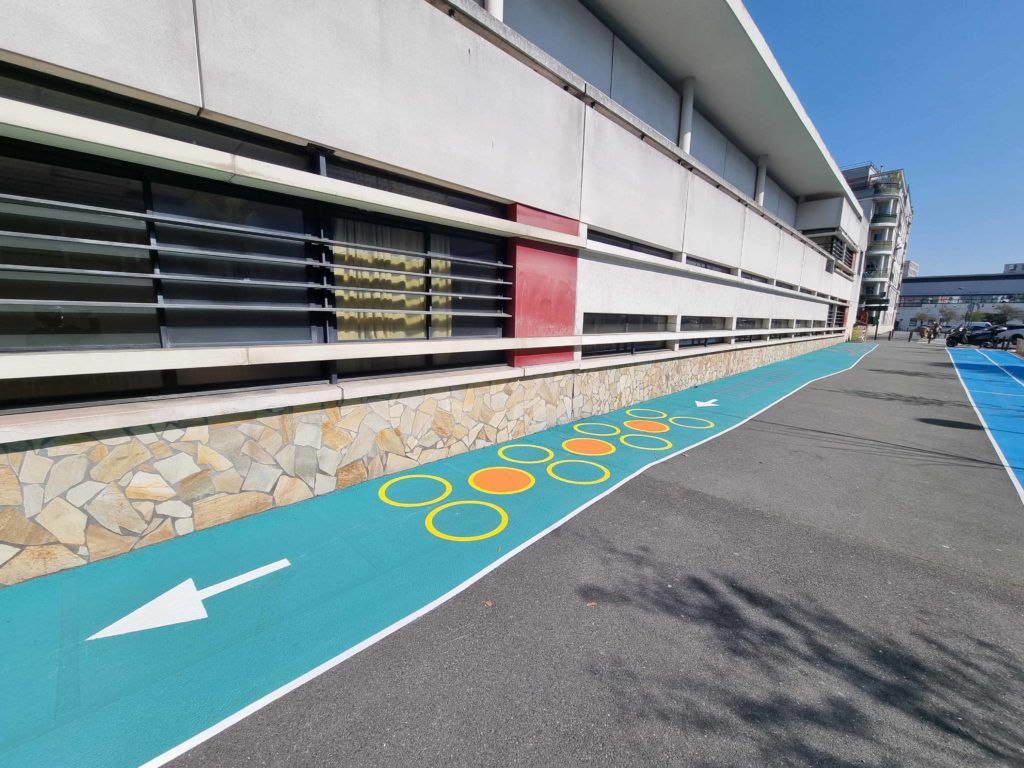 Parcours de motricité turquoise avec cercles jaunes oranges et flèche directionnelle devant le parvis de l'école